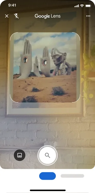 Sur un téléphone, l'application Google Lens est ouverte. Une notification indique à l'utilisateur qu'il peut appuyer sur le bouton de l'obturateur pour rechercher une image de grandes sculptures de mains en pierre dans un désert.